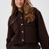 A woman wearing a dark brown button-up jacket with buttons and flap chest pockets, paired with dark brown pants, against a light gray studio background.