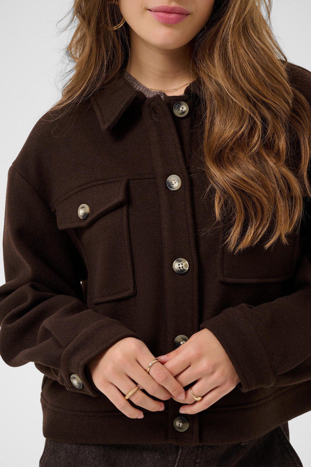 A close-up of a woman wearing a dark brown jacket with buttons and flap chest pockets, shown against a light gray studio background.