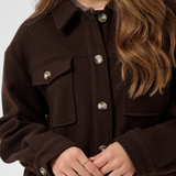 A close-up of a woman wearing a dark brown jacket with buttons and flap chest pockets, shown against a light gray studio background.