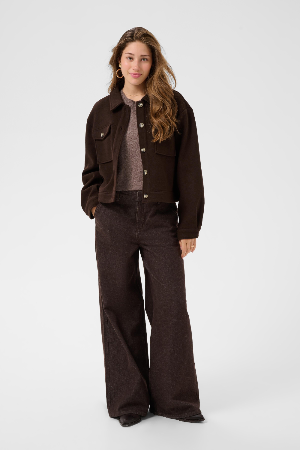 A woman modeling a dark brown cropped jacket and wide-leg pants over a  knit top, photographed against a light gray studio background.