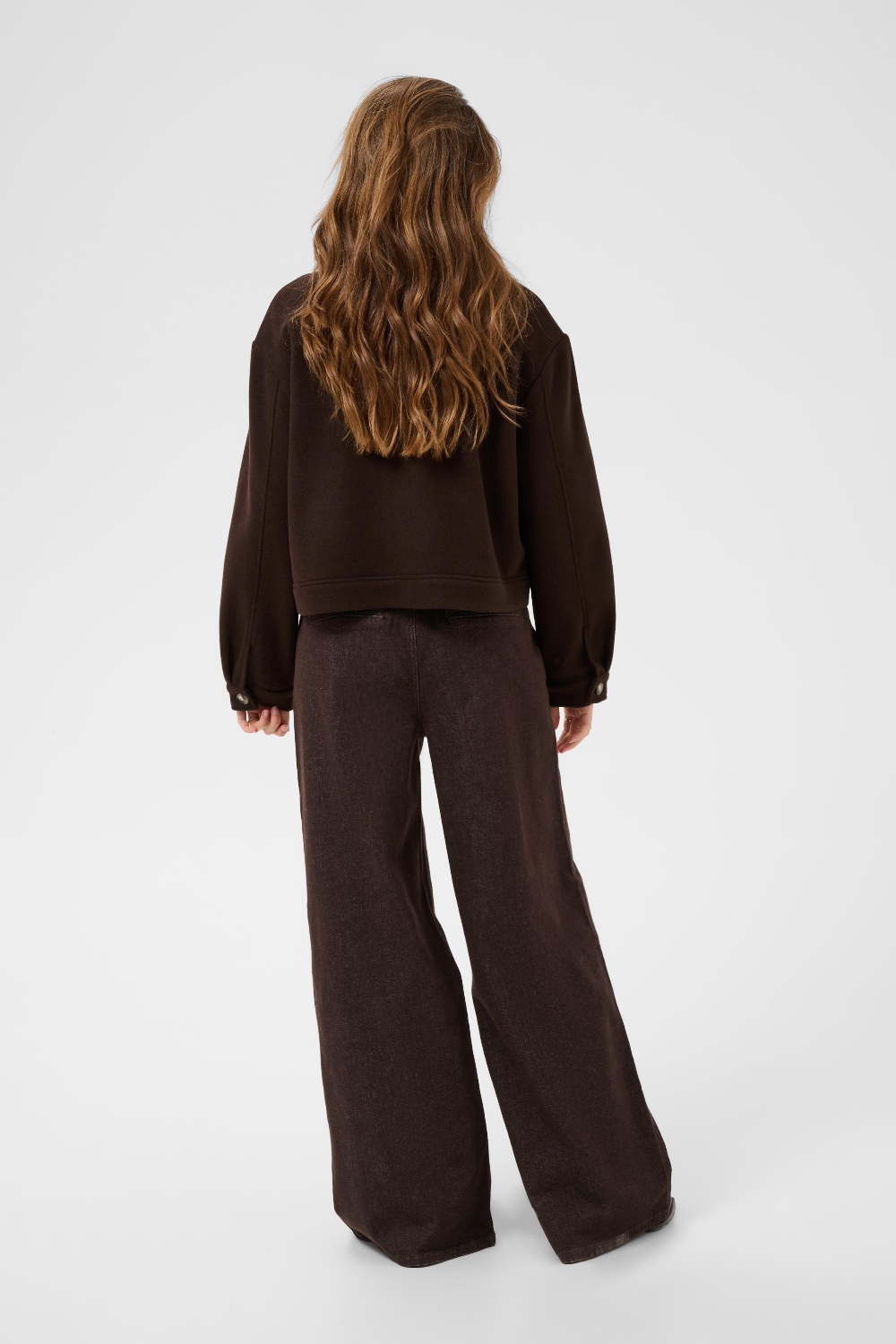 A model viewed from the back wearing a dark brown jacket and dark brown wide-leg trousers, set against a plain light gray studio background.