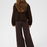 A model viewed from the back wearing a dark brown jacket and dark brown wide-leg trousers, set against a plain light gray studio background.