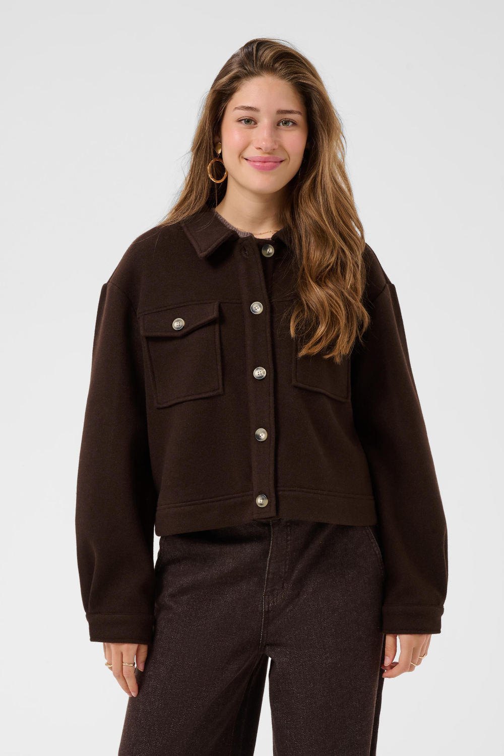 A woman modeling a dark brown button-up jacket with flap chest pockets, paired with dark brown pants, against a light gray studio background.