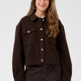 A woman modeling a dark brown button-up jacket with flap chest pockets, paired with dark brown pants, against a light gray studio background.