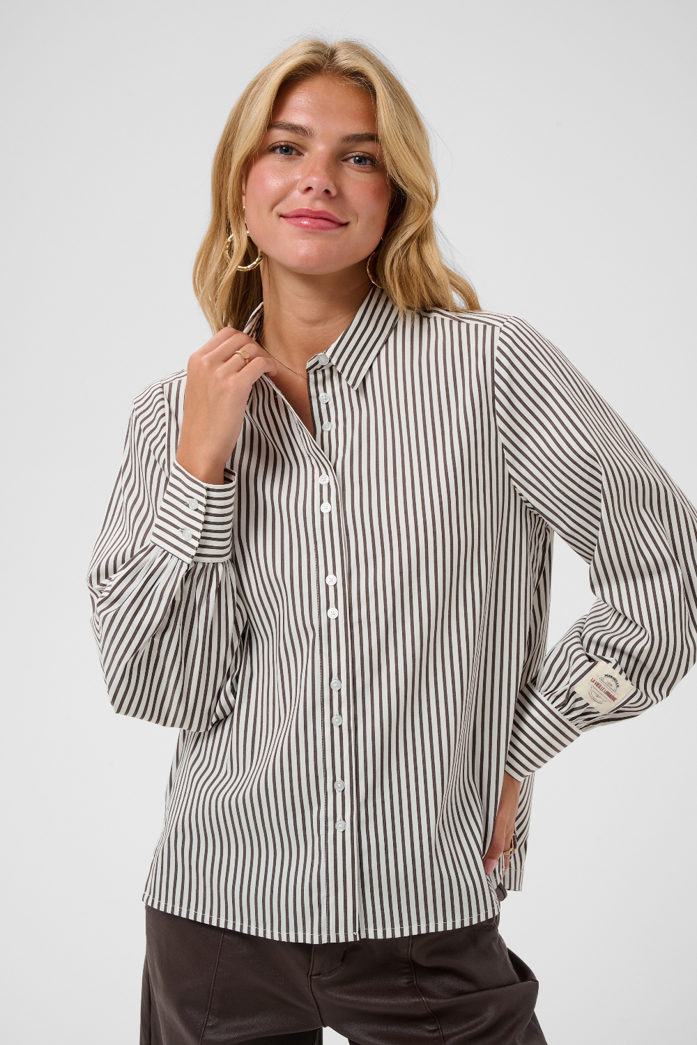 A woman  wearing a brown and white vertically striped long-sleeve blouse, paired with dark brown pants, against a light gray studio background.
