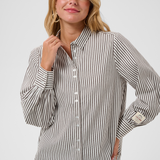 A woman  wearing a brown and white vertically striped long-sleeve blouse, paired with dark brown pants, against a light gray studio background.