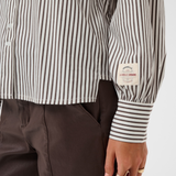 Close-up of a person wearing a brown and white striped shirt with a visible brand label, standing against a white background.