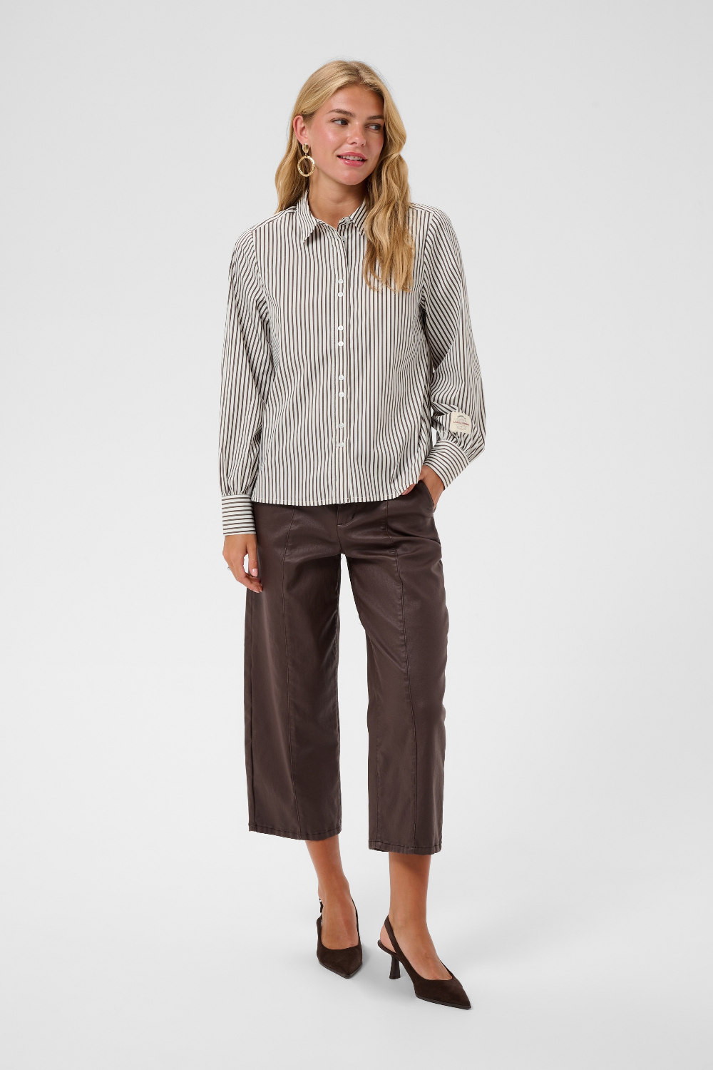 A woman  wearing a brown and white vertically striped long-sleeve blouse, paired with dark brown cropped pants and black sandals, against a light gray studio background.