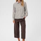 A woman  wearing a brown and white vertically striped long-sleeve blouse, paired with dark brown cropped pants and black sandals, against a light gray studio background.