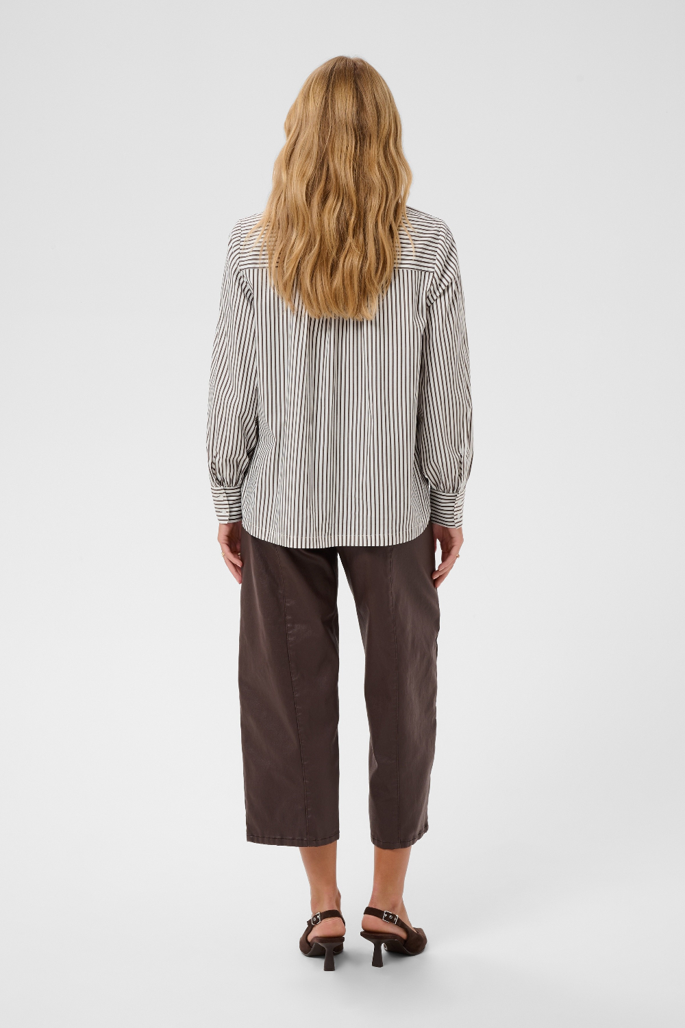 A woman viewed from behind wearing a brown and white vertically striped long-sleeve blouse, paired with dark brown cropped pants and black sandals, against a light gray studio background.