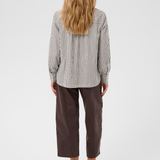 A woman viewed from behind wearing a brown and white vertically striped long-sleeve blouse, paired with dark brown cropped pants and black sandals, against a light gray studio background.
