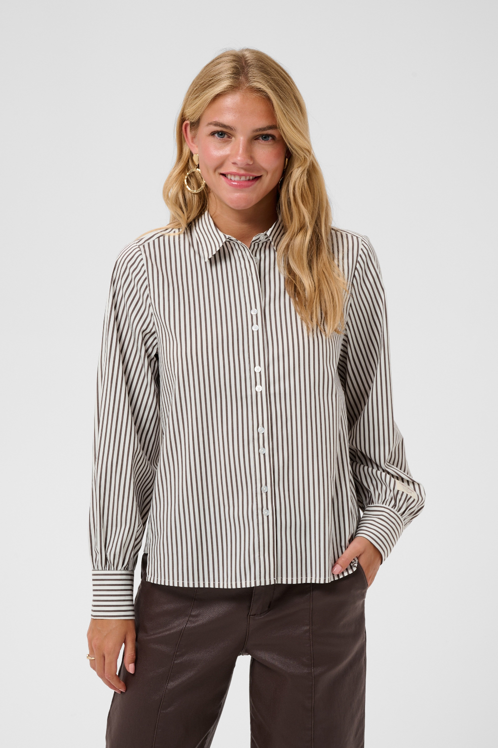 A woman  wearing a brown and white vertically striped long-sleeve blouse, paired with dark brown pants, against a light gray studio background.