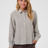 A woman  wearing a brown and white vertically striped long-sleeve blouse, paired with dark brown pants, against a light gray studio background.