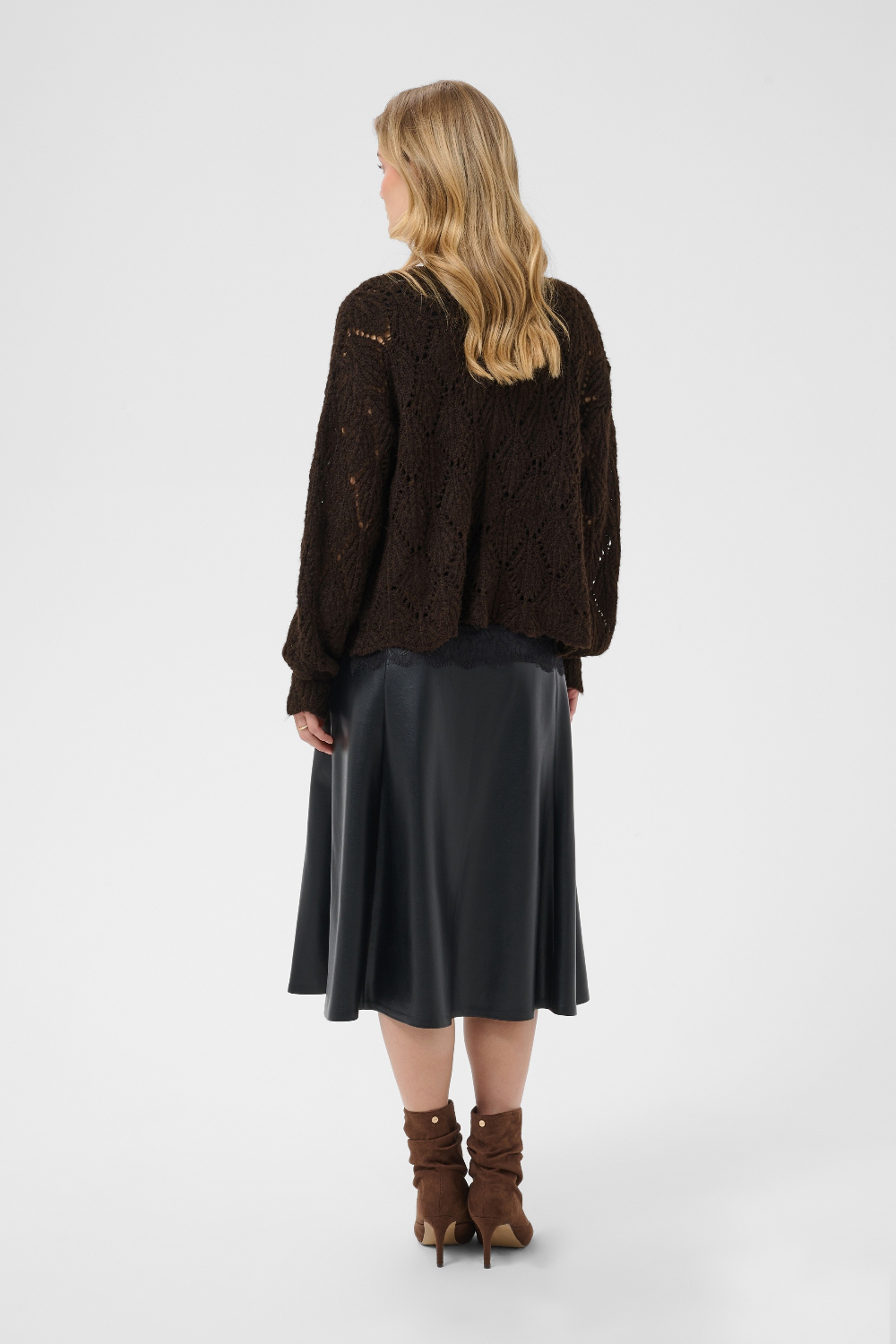 A woman is shown from the back wearing a dark brown open-knit sweater, a black satin midi skirt, and brown heeled ankle boots against a light gray studio background.