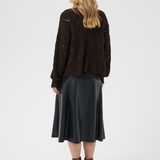 A woman is shown from the back wearing a dark brown open-knit sweater, a black satin midi skirt, and brown heeled ankle boots against a light gray studio background.