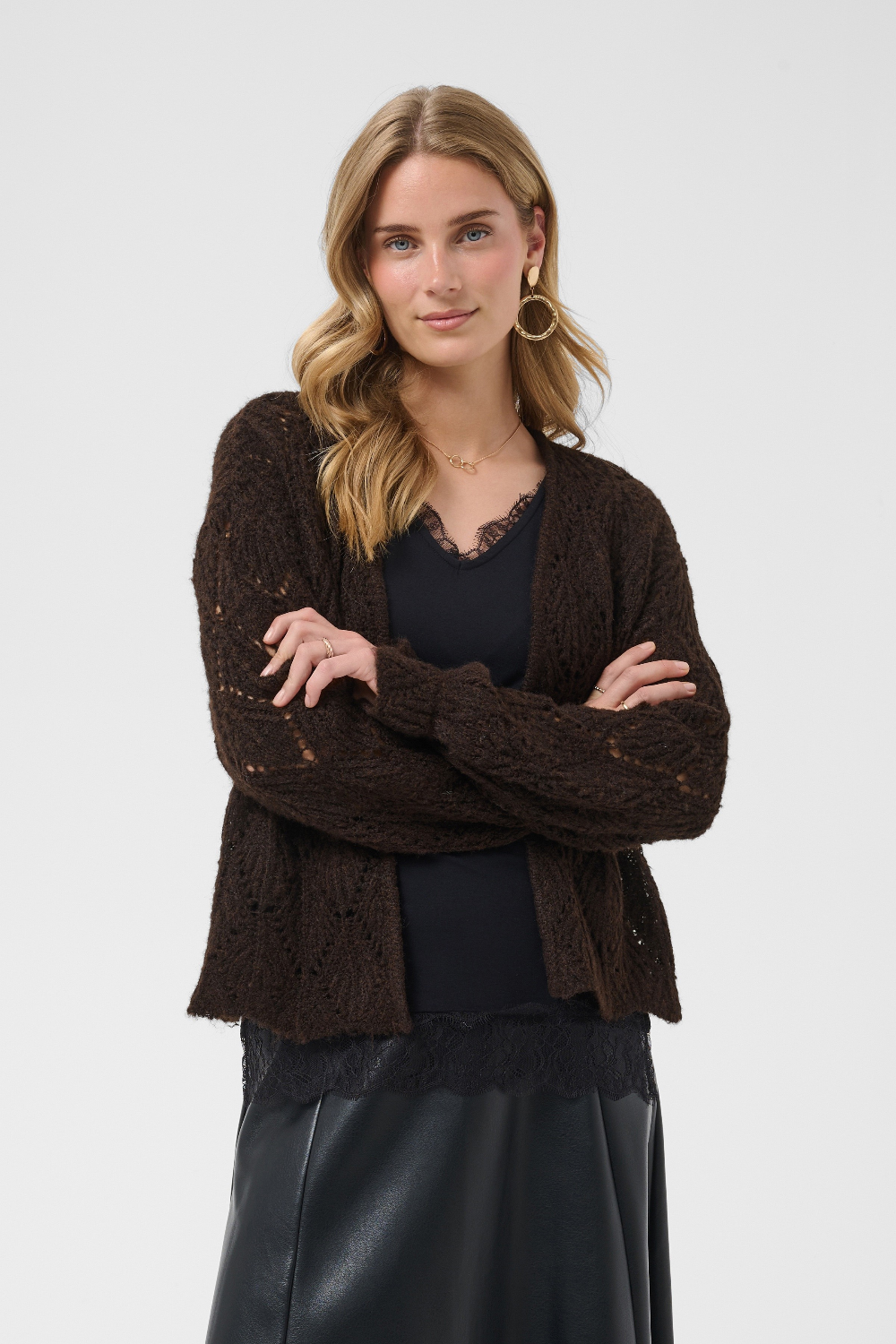 A woman modeling a dark brown open-front knit cardigan with a textured pattern, worn over a black top and black skirt against a light gray studio background.