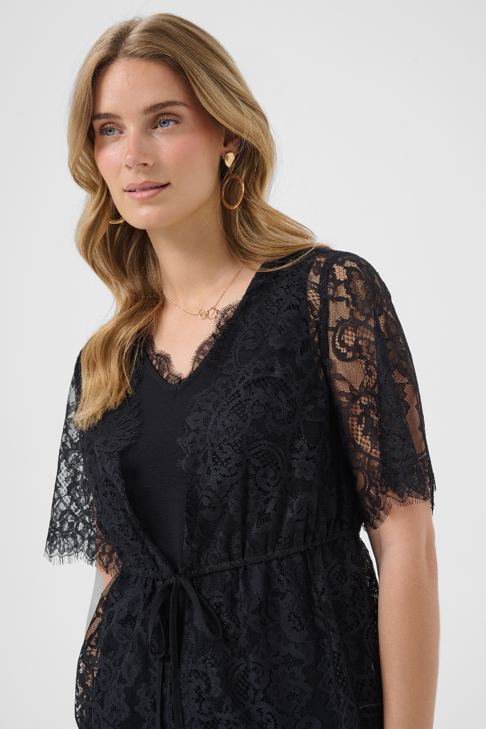 A woman wearing a black lace top with short sleeves and a tie waist, photographed against a light gray studio background.