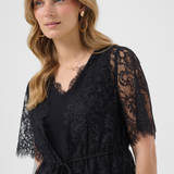 A woman wearing a black lace top with short sleeves and a tie waist, photographed against a light gray studio background.