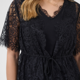 A close-up of a woman wearing a black lace top with short sleeves, a V-neckline, and a drawstring waist, shown against a light gray studio background.