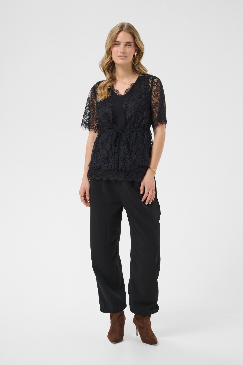 A woman modeling a black lace short-sleeve top with a tie waist, paired with black wide-leg pants and brown ankle boots, against a light gray studio background.