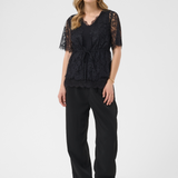 A woman modeling a black lace short-sleeve top with a tie waist, paired with black wide-leg pants and brown ankle boots, against a light gray studio background.