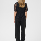 A woman viewed from behind wearing a black lace short-sleeve top and black wide-leg trousers, standing against a light gray studio background.