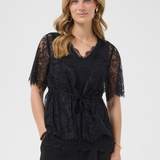 A woman modeling a black lace short-sleeve top with a tie waist, paired with black pants, against a light gray studio background.