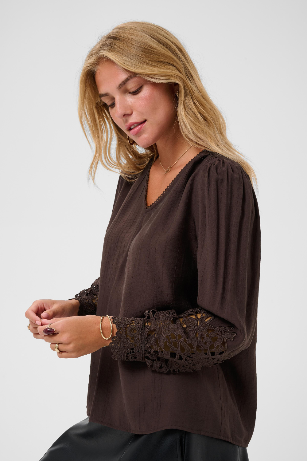 A woman wearing a dark brown long-sleeve blouse with intricate lace detailing on the sleeves, shown against a plain light gray studio background.