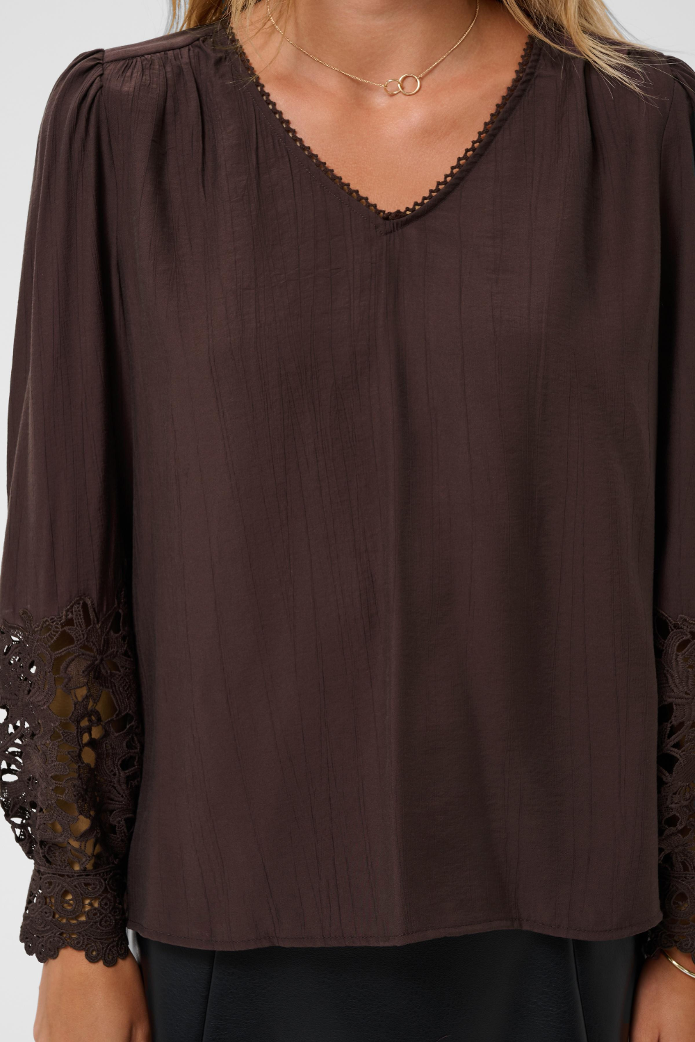 A woman wearing a dark brown long-sleeve blouse with a V-neckline, gathered shoulders, and black lace trim at the cuffs and neckline.