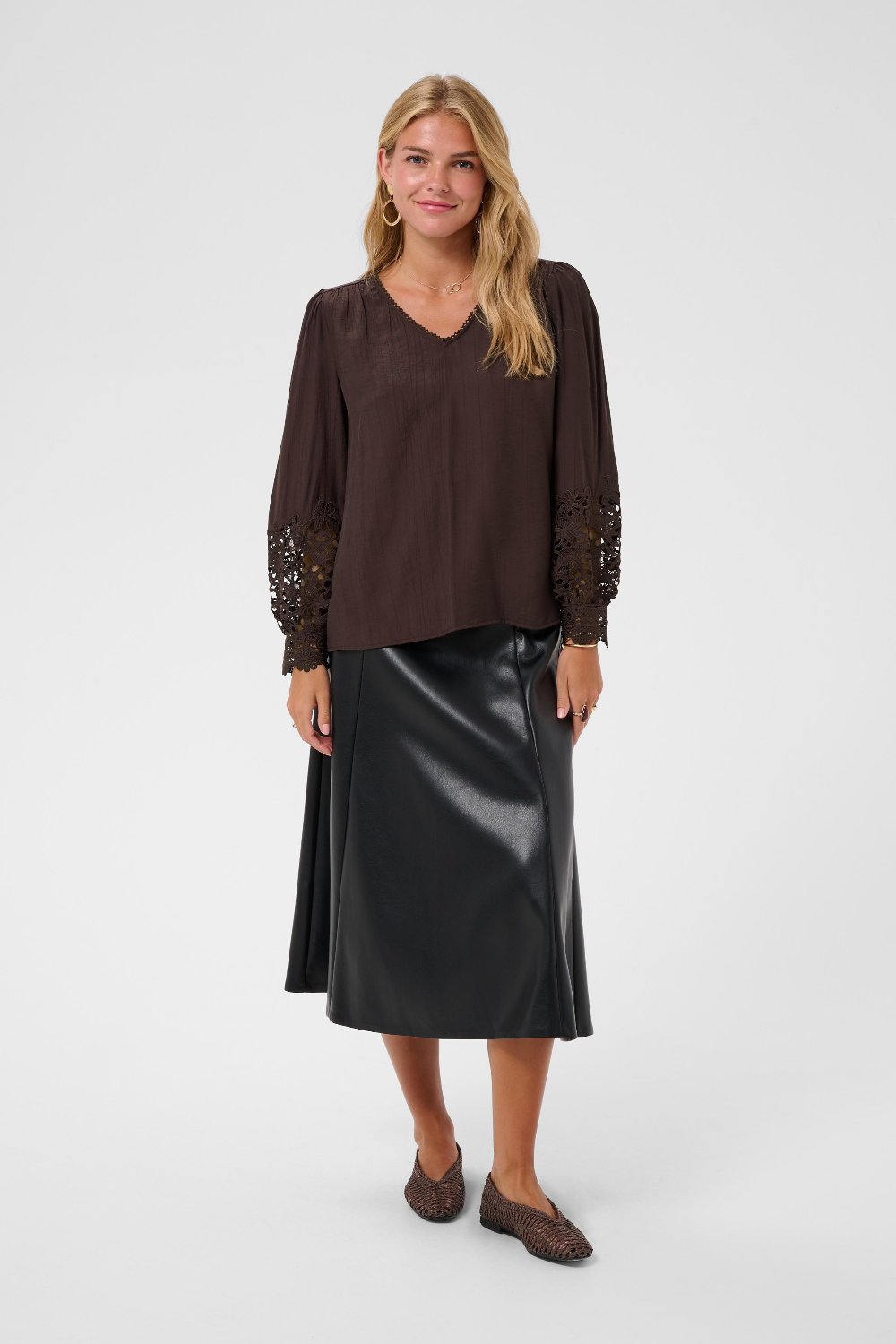 A woman modeling a dark brown long-sleeve blouse with lace sleeve cuffs and a black faux-leather midi skirt, paired with brown flat shoes, against a light gray studio background.