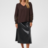 A woman modeling a dark brown long-sleeve blouse with lace sleeve cuffs and a black faux-leather midi skirt, paired with brown flat shoes, against a light gray studio background.