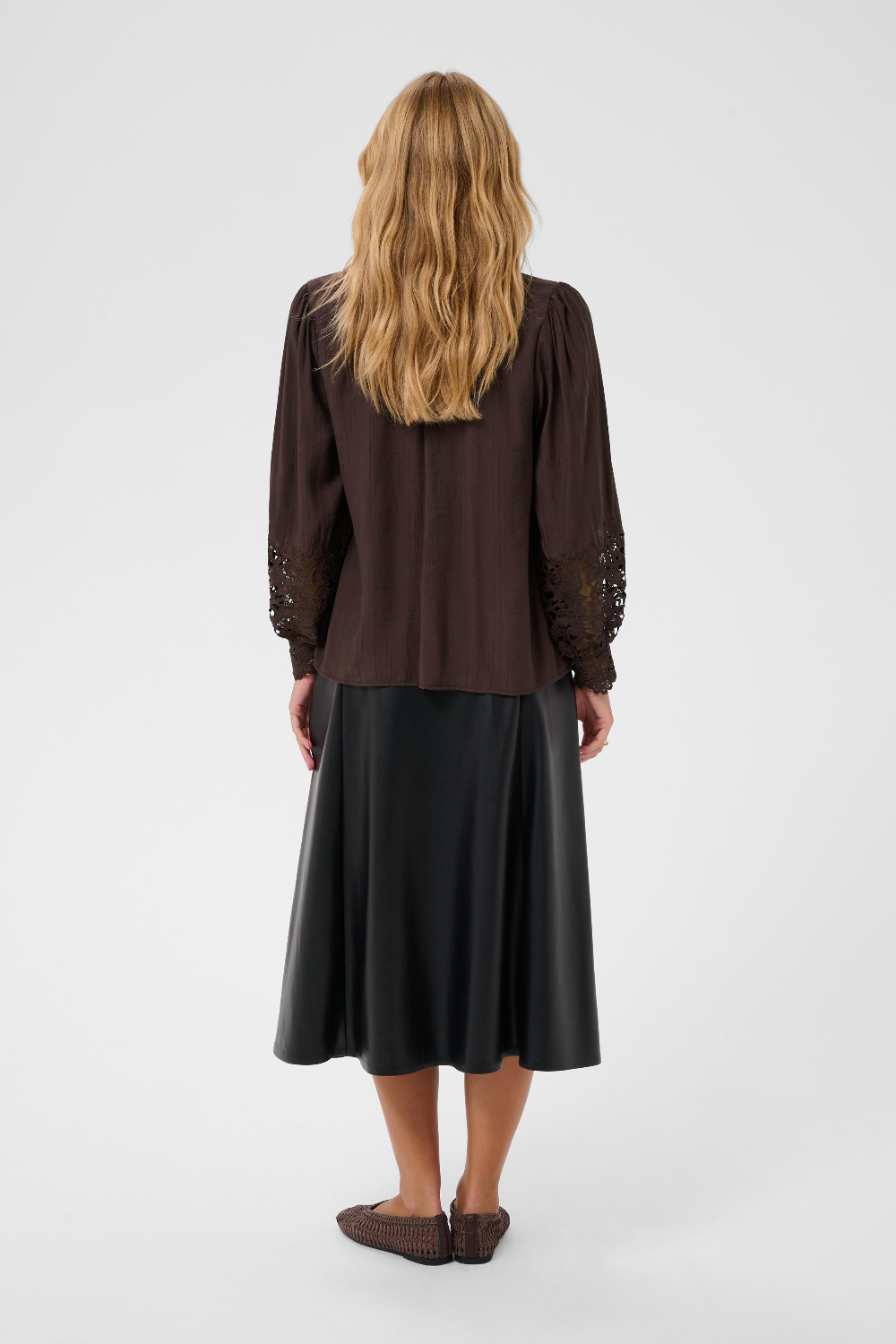A woman viewed from the back wearing a dark brown long-sleeve blouse with lace sleeve detailing and a black midi skirt, standing against a light gray studio background.