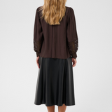 A woman viewed from the back wearing a dark brown long-sleeve blouse with lace sleeve detailing and a black midi skirt, standing against a light gray studio background.