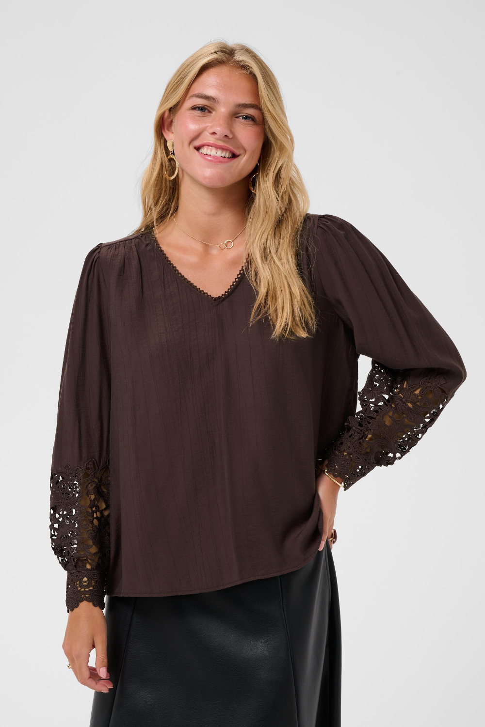 A woman wearing a dark brown long-sleeve blouse with sheer lace sleeves and a V-neckline, paired with a black skirt, against a light gray studio background.
