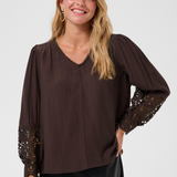 A woman wearing a dark brown long-sleeve blouse with sheer lace sleeves and a V-neckline, paired with a black skirt, against a light gray studio background.
