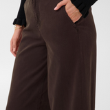 Close up of a woman modelling a black sweater and a brown trouser style denim, with her hand in the pocket, against a light backdrop.