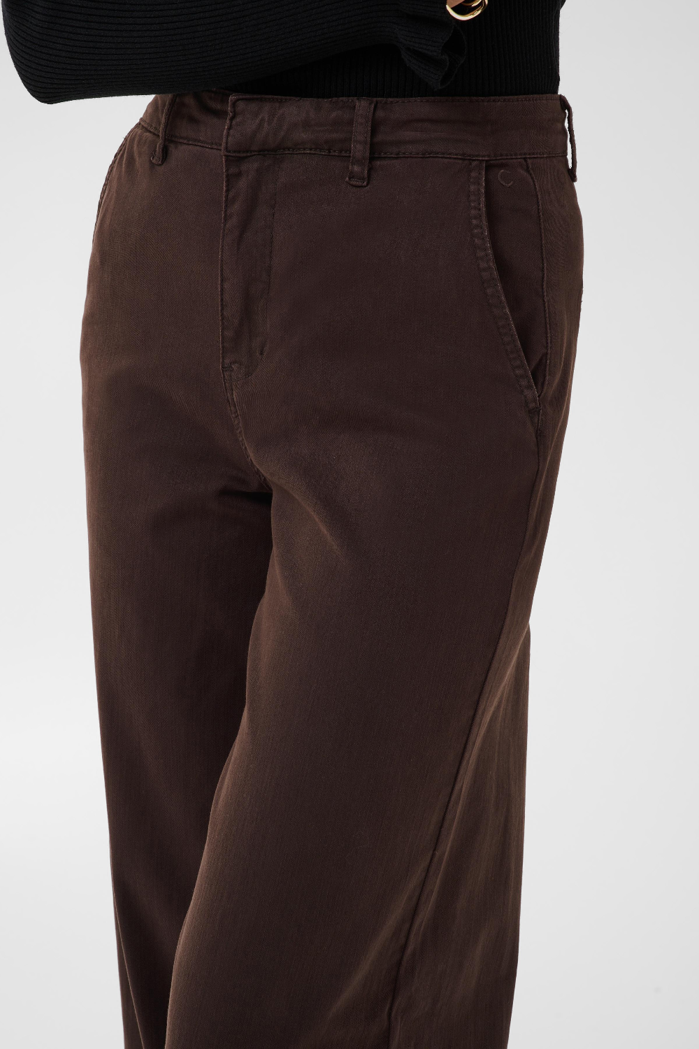 Close up of a person modelling a black sweater and a brown trouser style denim against a light backdrop.