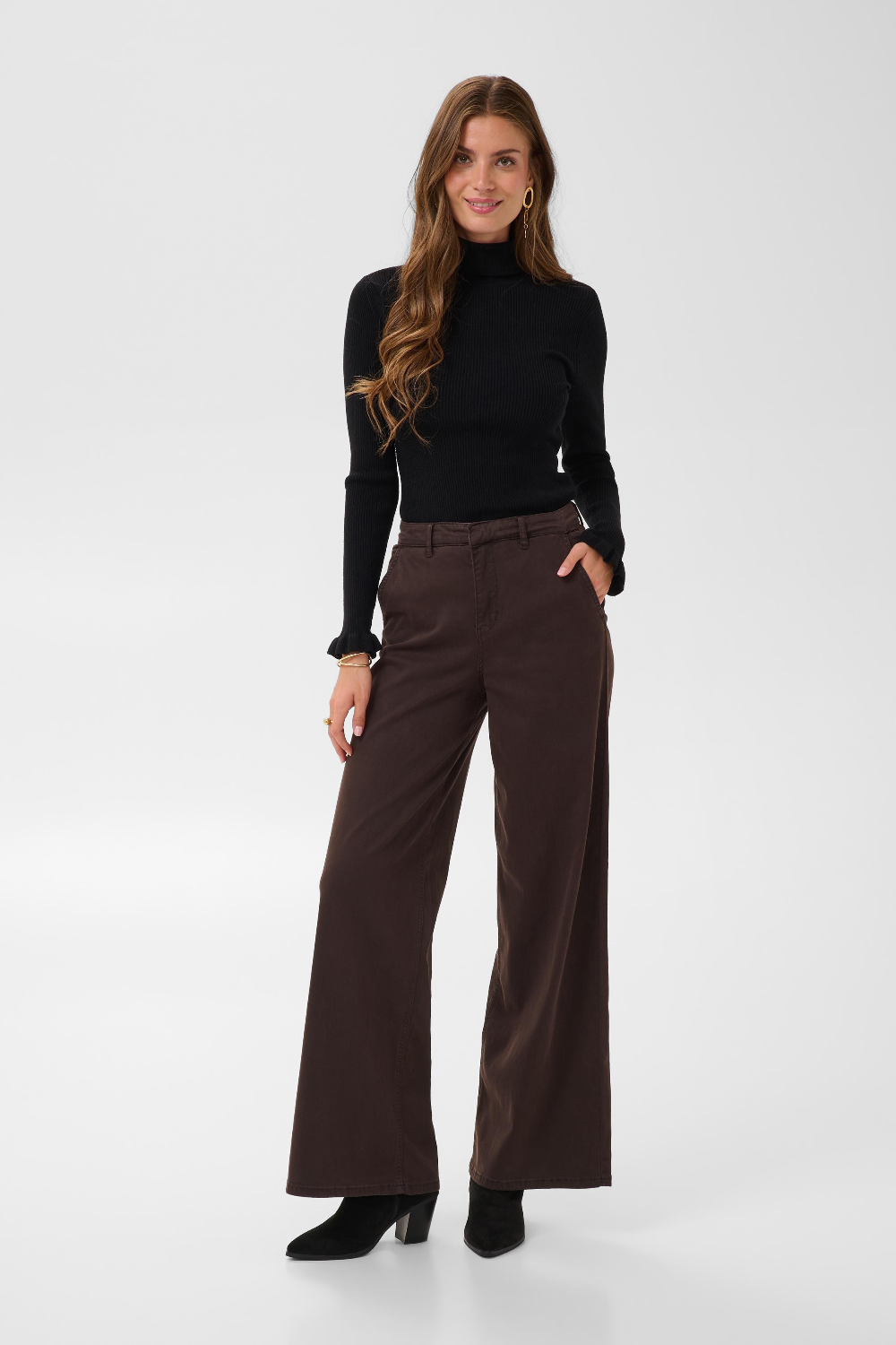 Woman with long hair modelling a black turtle neck sweater and a wide leg brown trouser style denim with black high heel boots against a light backdrop.