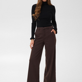 Woman with long hair modelling a black turtle neck sweater and a wide leg brown trouser style denim with black high heel boots against a light backdrop.