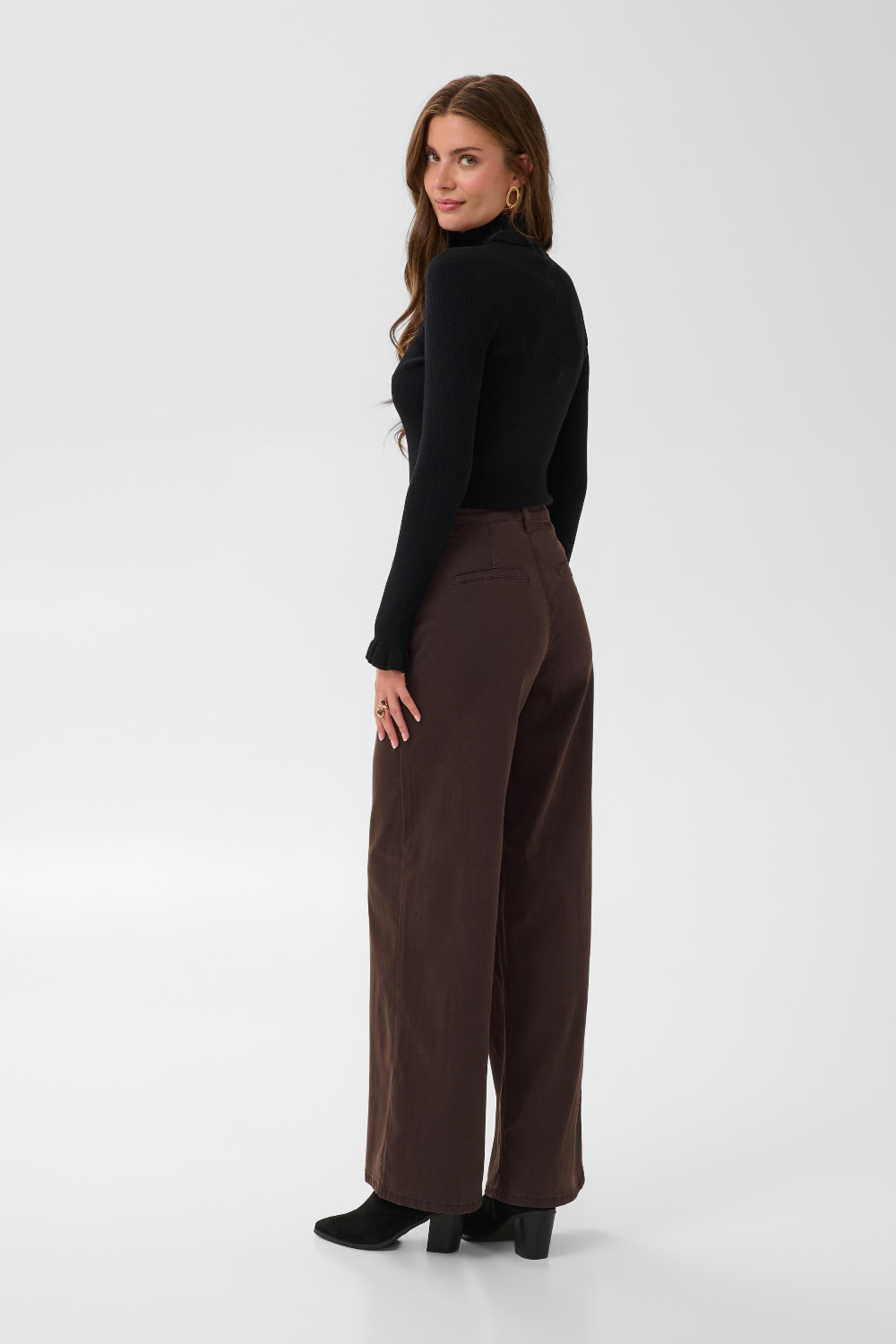 Woman with long hair, shown from behind, modelling a black turtle neck sweater and a wide leg brown trouser style denim with black high heel boots against a light backdrop.