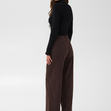 Woman with long hair, shown from behind, modelling a black turtle neck sweater and a wide leg brown trouser style denim with black high heel boots against a light backdrop.