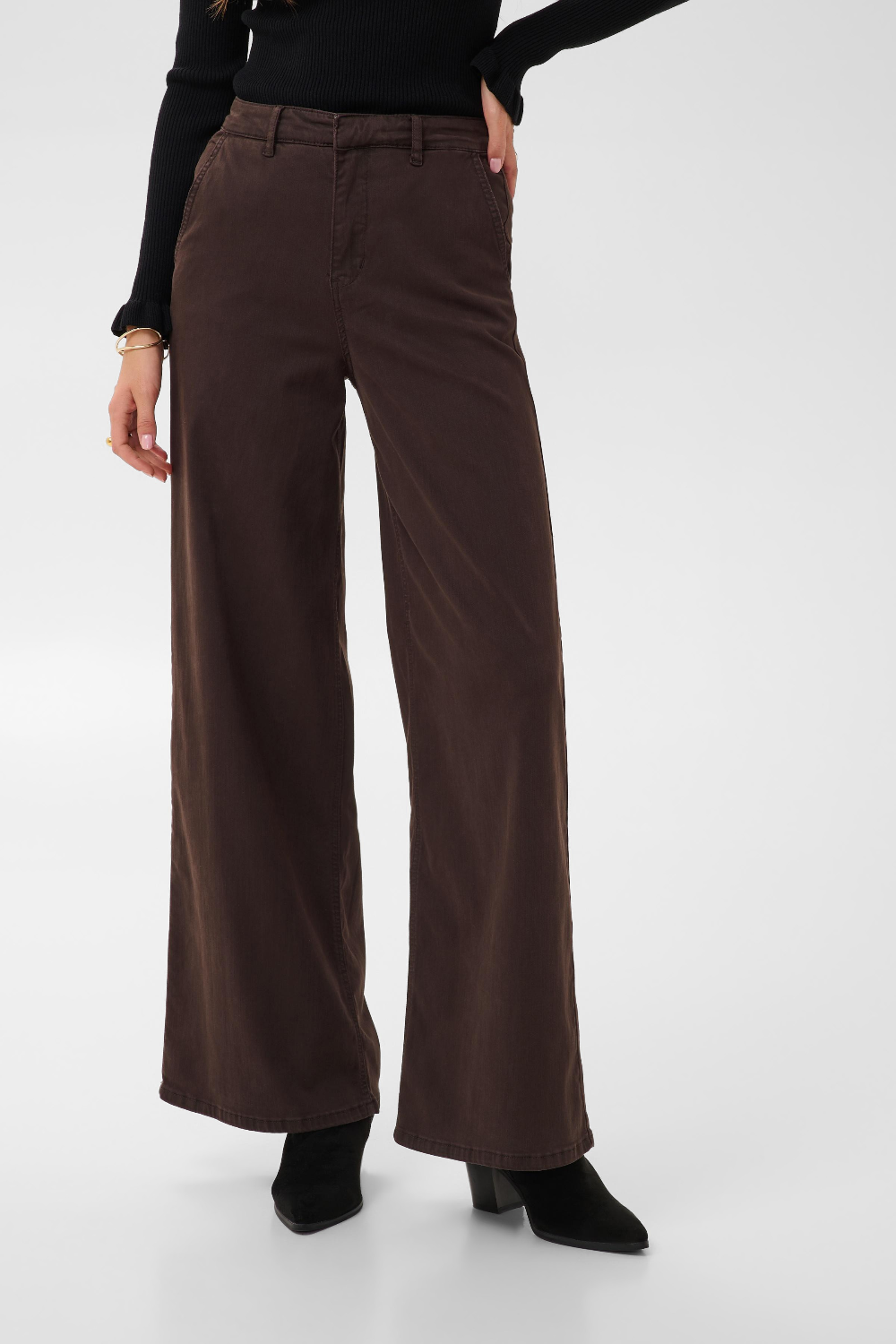 Woman modelling a black sweater and a wide leg brown trouser style denim with black high heel boots against a light backdrop.