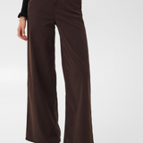 Woman modelling a black sweater and a wide leg brown trouser style denim with black high heel boots against a light backdrop.
