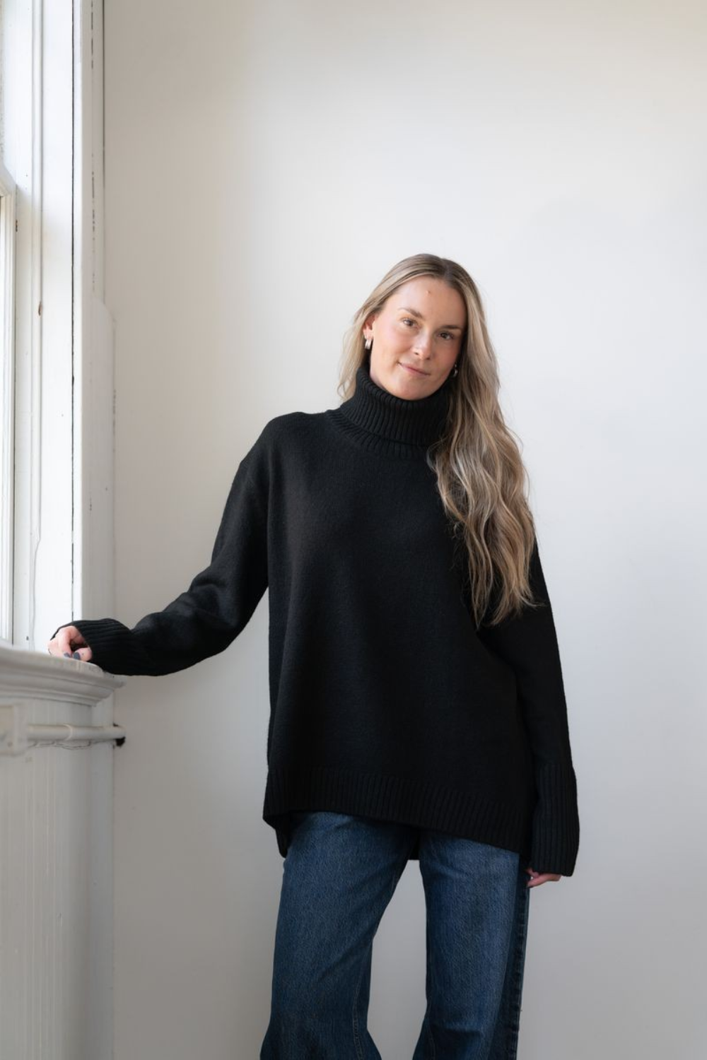 Woman modelling an oversized black cowl neck sweater with blue jeans in a white room photographed near a window