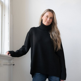Woman modelling an oversized black cowl neck sweater with blue jeans in a white room photographed near a window