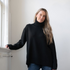 Woman modelling an oversized black cowl neck sweater with blue jeans in a white room photographed near a window