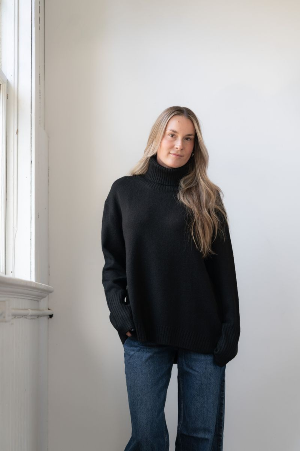 Woman modelling an oversized black cowl neck sweater with blue jeans in a white room photographed near a window