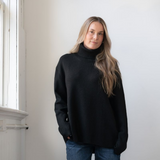 Woman modelling an oversized black cowl neck sweater with blue jeans in a white room photographed near a window
