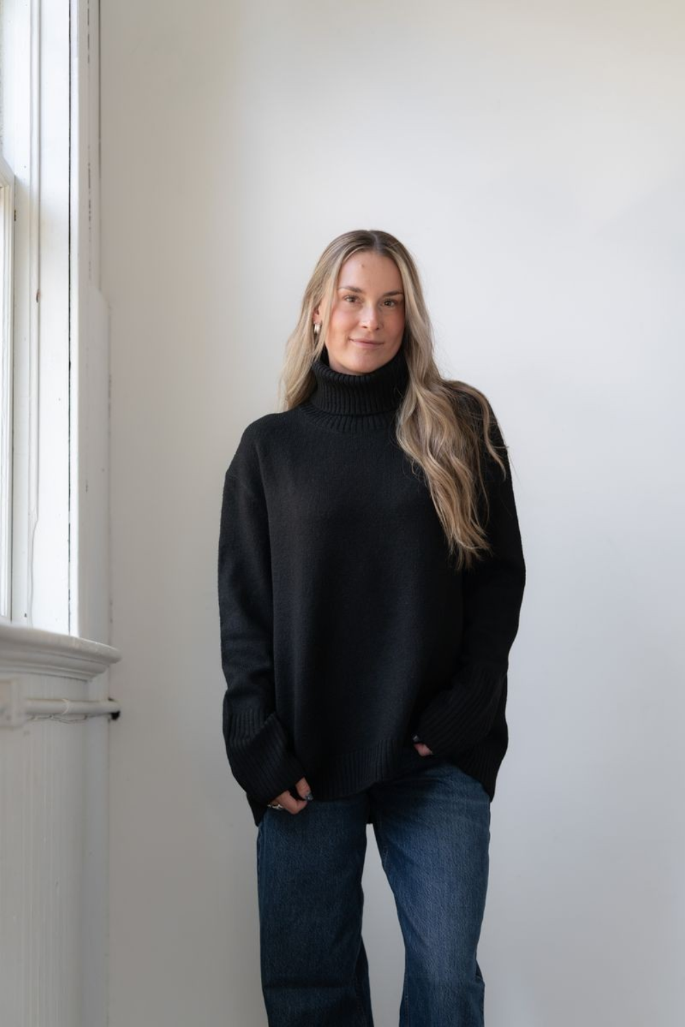 Woman modelling an oversized black cowl neck sweater with blue jeans in a white room photographed near a window