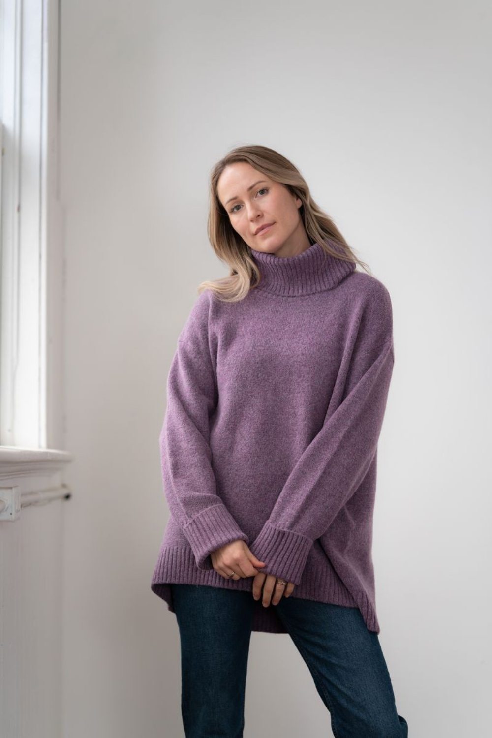 Woman modelling an oversized purple cowl neck sweater with blue jeans in a white room photographed near a window
